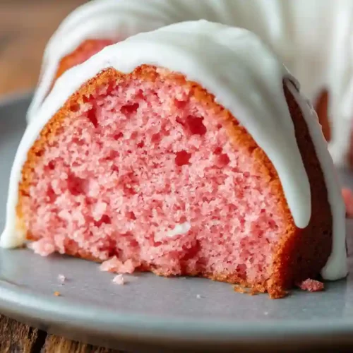 Slice of strawberry bundt cake with uniform pink crumb and thick cream cheese glaze on a gray plate