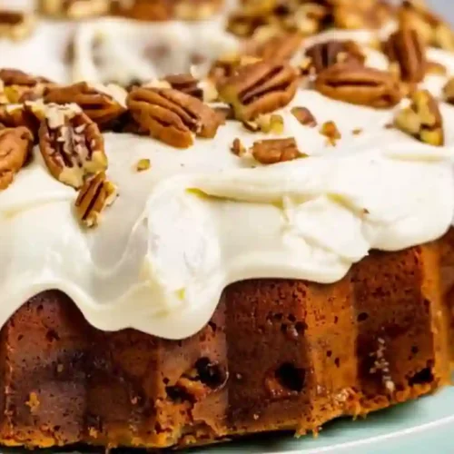 Close-up of a moist banana bundt cake topped with cream cheese frosting and chopped pecans on a white plate.