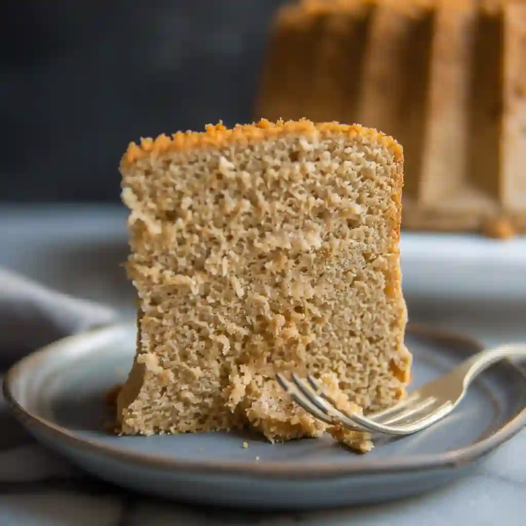 A slice of coffee chiffon cake on a small plate with a fork, showing airy texture and golden brown crust, whole cake blurred in background
