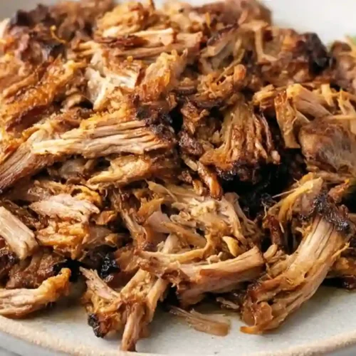 Close-up of crispy shredded pork carnitas served on a light gray ceramic plate with lime wedges and cilantro