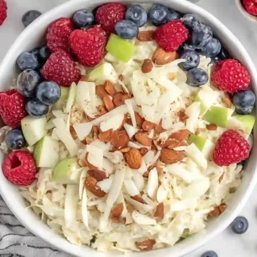Close-up of bircher muesli topped with raspberries, blueberries, green apple pieces, and almonds in a ceramic bowl