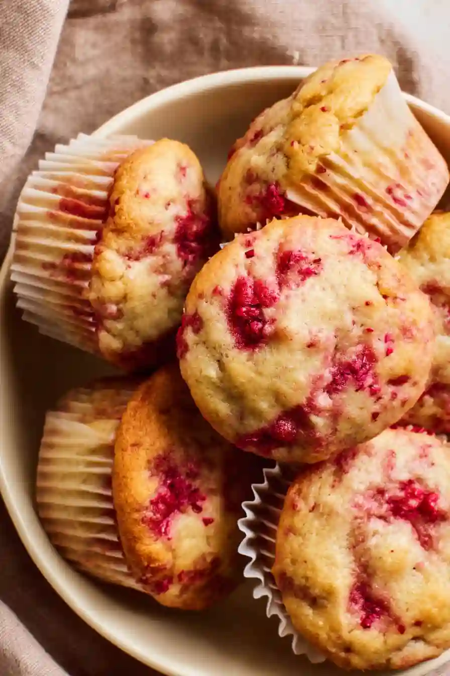 Vertical image of soft raspberry muffins with red berry swirls in white liners, photographed from above