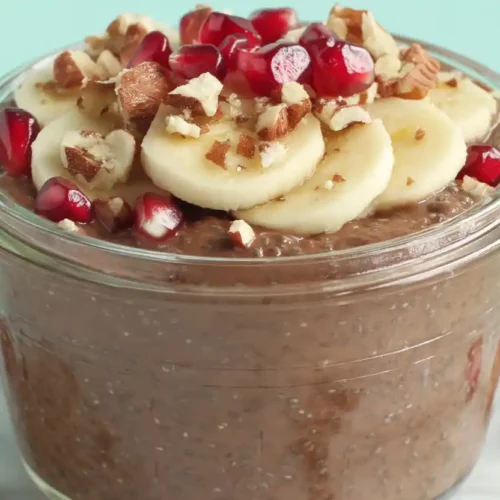 Chocolate chia pudding in a clear glass jar with visible chia seeds, topped with banana slices, nuts, and pomegranate seeds on a soft pastel background