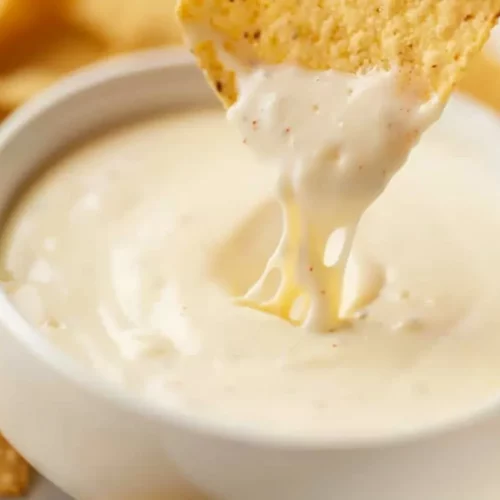 Extra close-up of creamy white cheese dip in a bowl with a tortilla chip dipping and melted cheese dripping down.