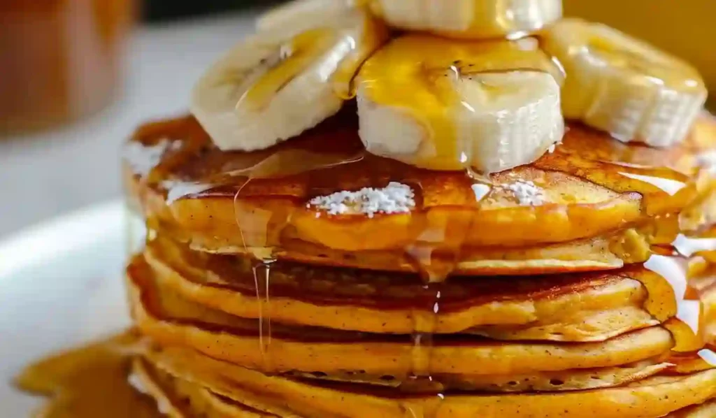 Stack of fluffy pumpkin pancakes topped with banana slices and maple syrup drizzle on a white plate