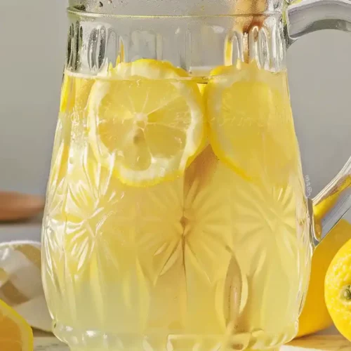 Close-up of homemade lemonade in an embossed glass pitcher with lemon slices on a light wooden surface.