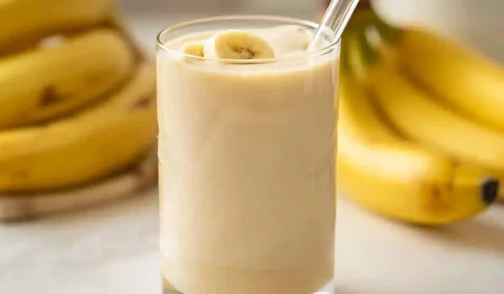 Creamy Korean banana milk in a clear glass with glass straw on marble countertop with bananas in background