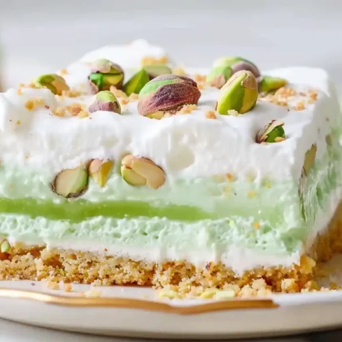 Close-up layered pistachio dessert bar with green filling, white cream topping, and pistachio garnish on a light plate