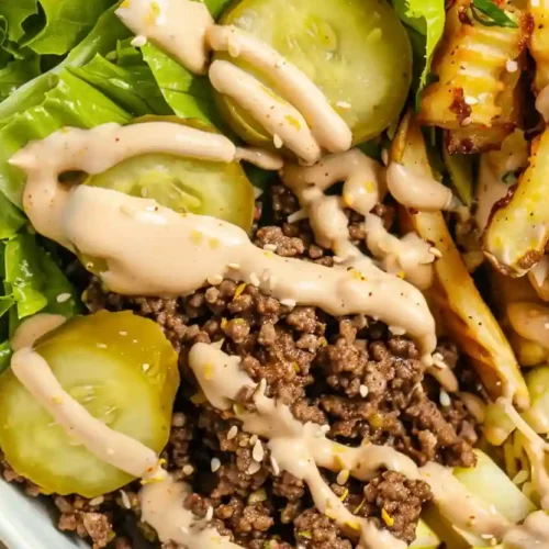 Overhead burger bowl with seasoned ground beef, romaine lettuce, pickles, shredded cheese and crispy fries drizzled with sauce