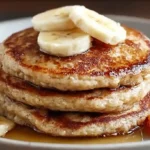Close-up stack of banana oatmeal pancakes topped with banana slices, syrup, and strawberries